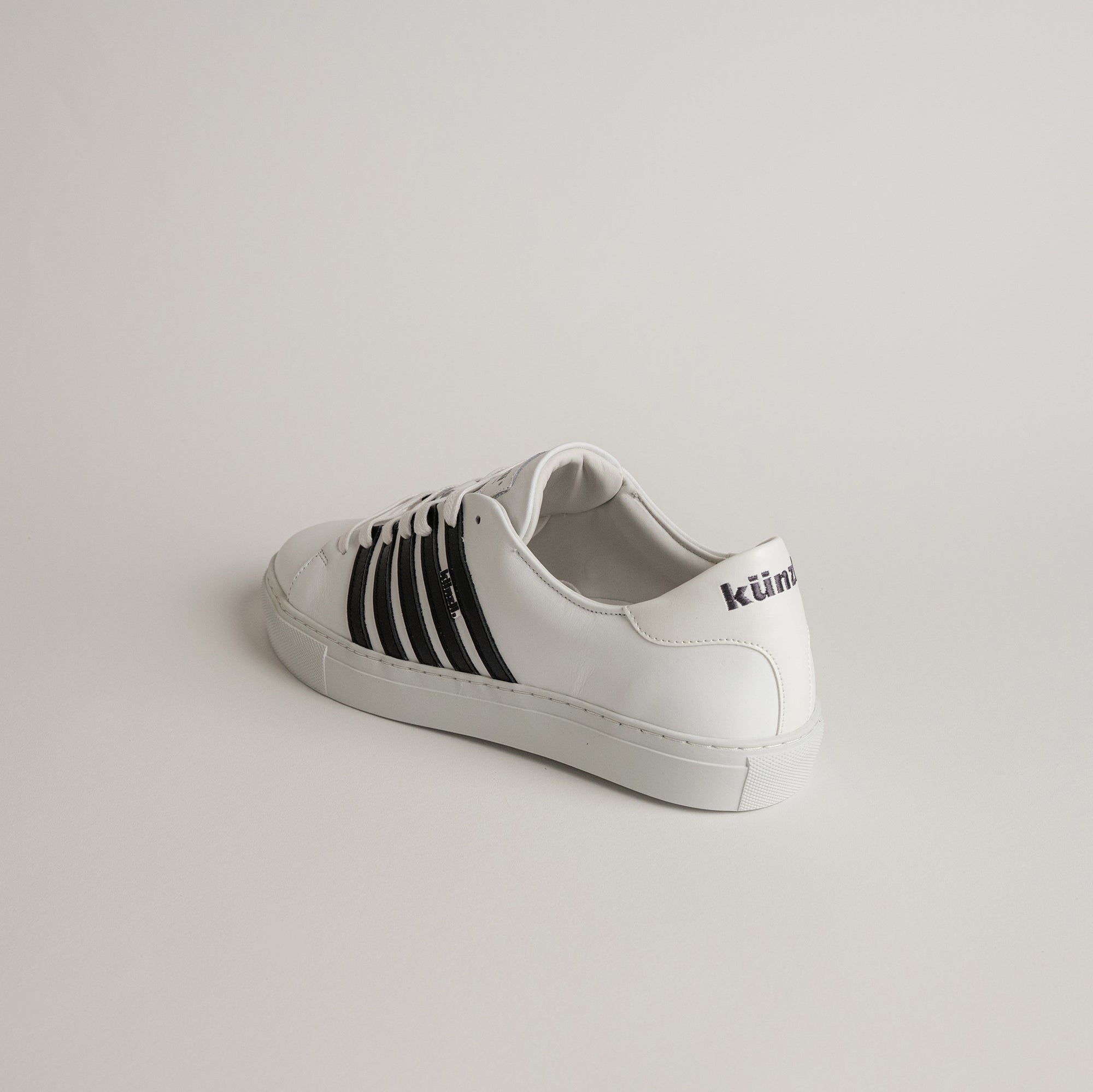 Weißer Sneaker von Kuenzli Schuhe mit schwarzen Streifen auf hellem Hintergrund.