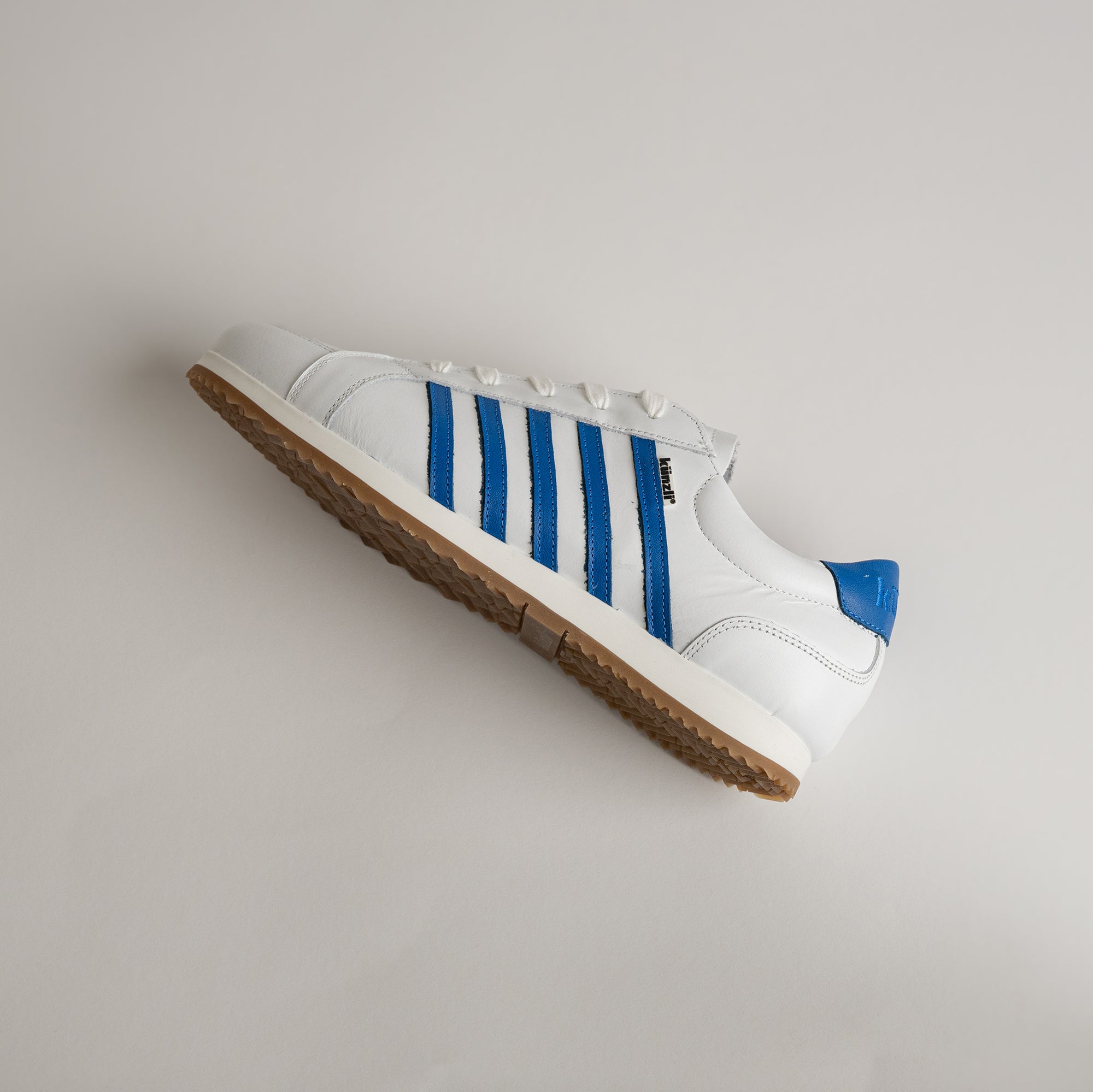 Weißer Kuenzli Schuh mit blauen Streifen auf grauem Hintergrund, sportlich und modern.