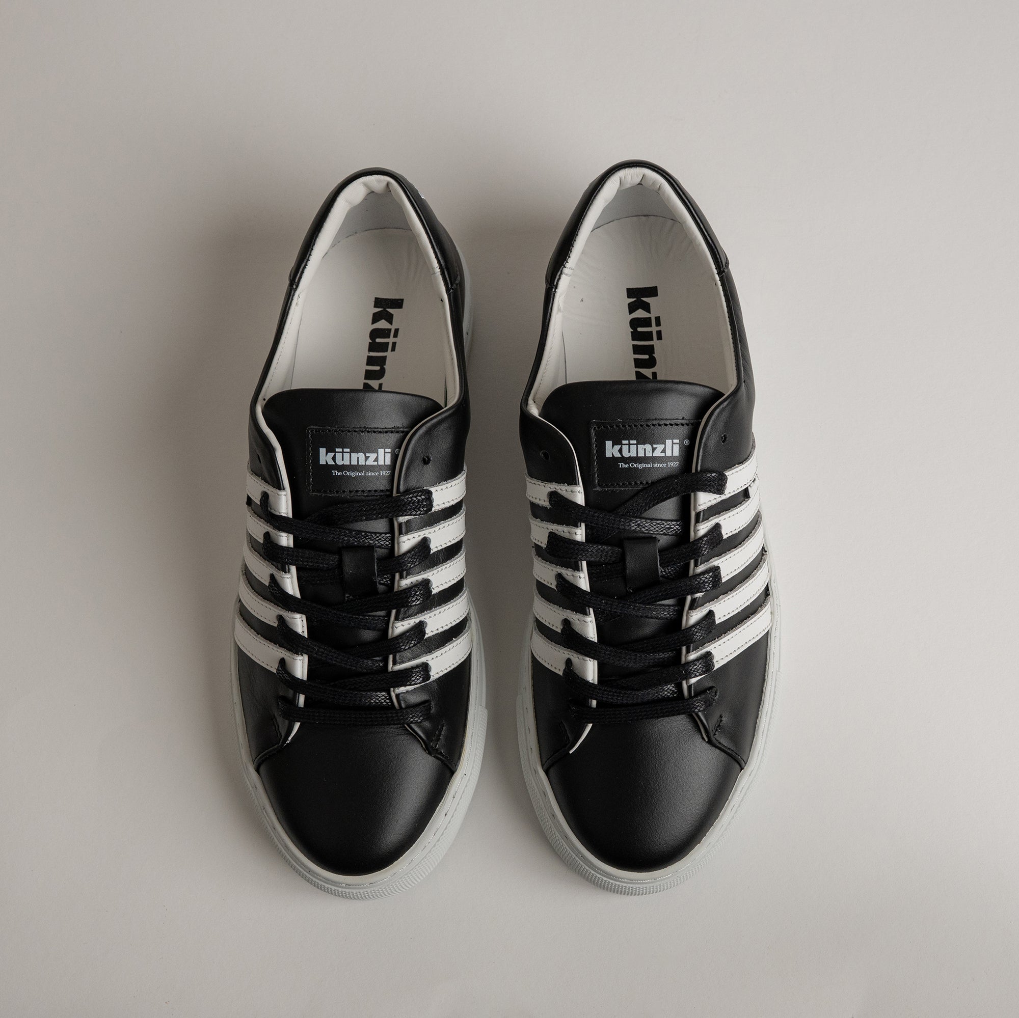 Schwarze Kuenzli Schuhe mit weißen Streifen, aus Leder, auf hellem Hintergrund.