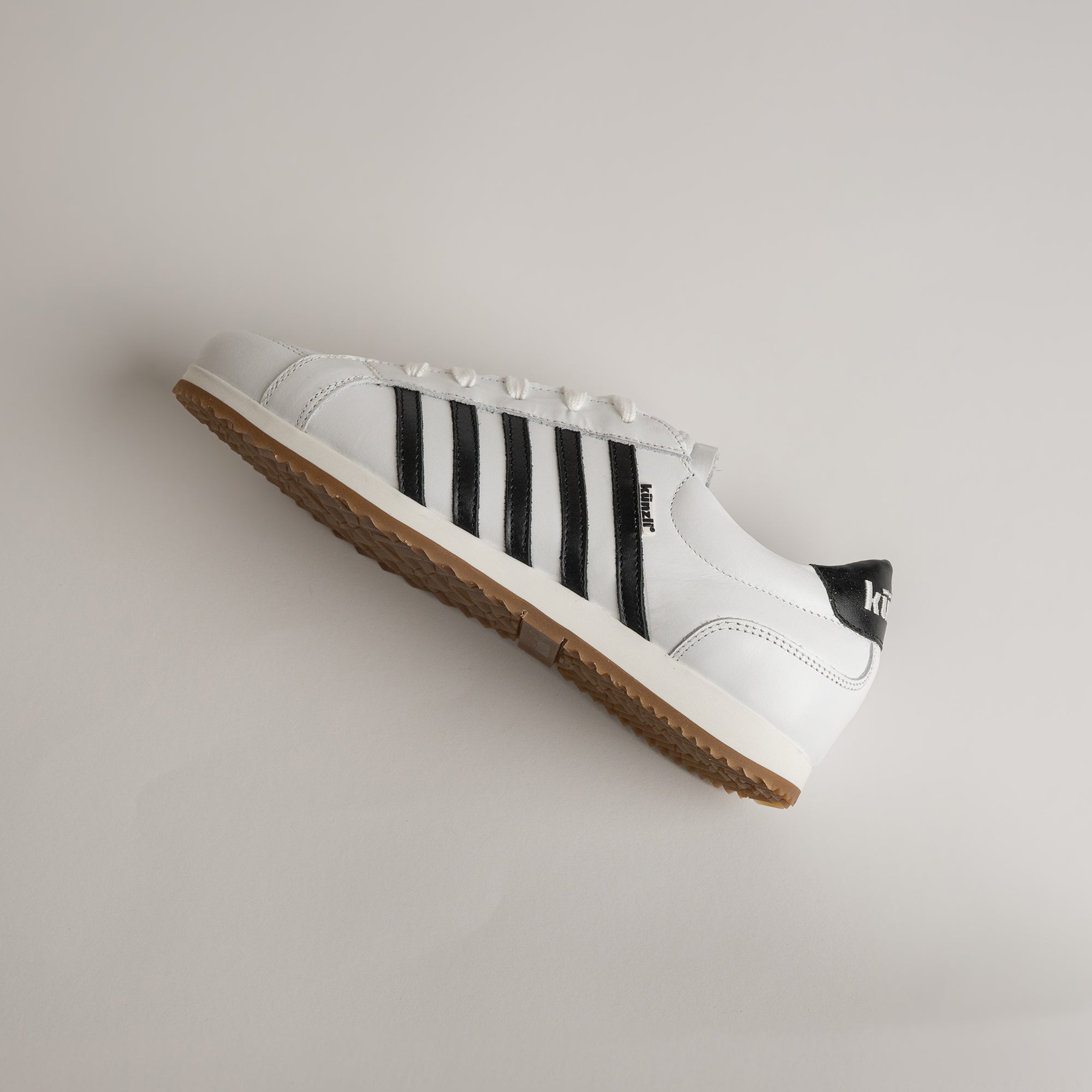 Weißer Kuenzli Schuh mit schwarzen Streifen und brauner Sohle auf grauem Hintergrund.