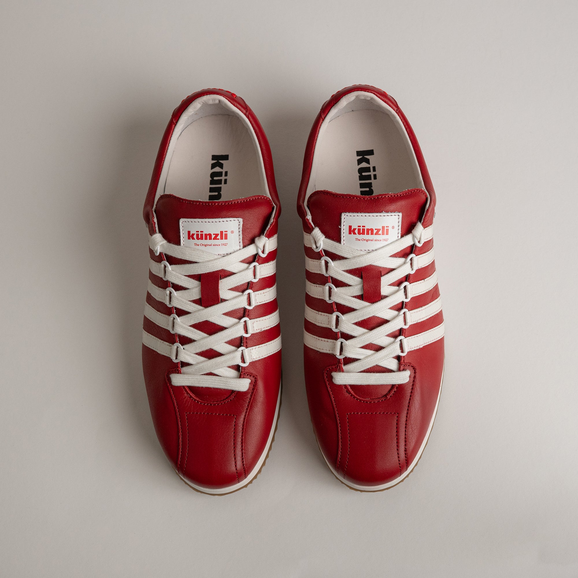 Rote Kuenzli Schuhe aus Leder mit weißen Streifen auf grauem Hintergrund.