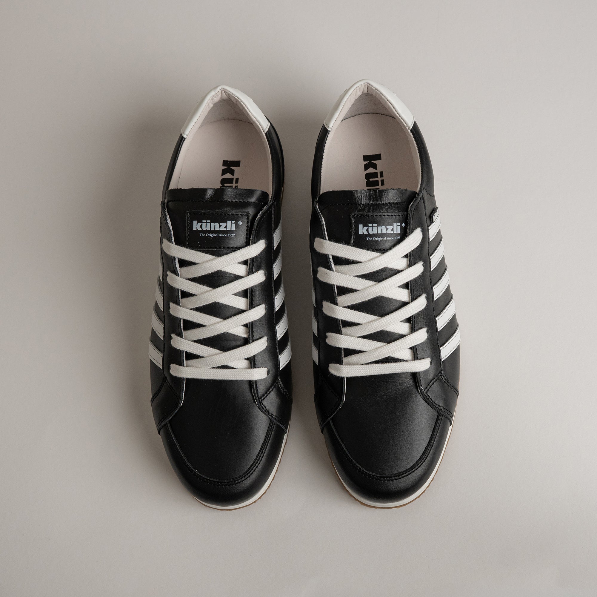 Schwarze Kuenzli Schuhe aus Leder mit weißen Streifen und Schnürsenkeln auf grauem Hintergrund.