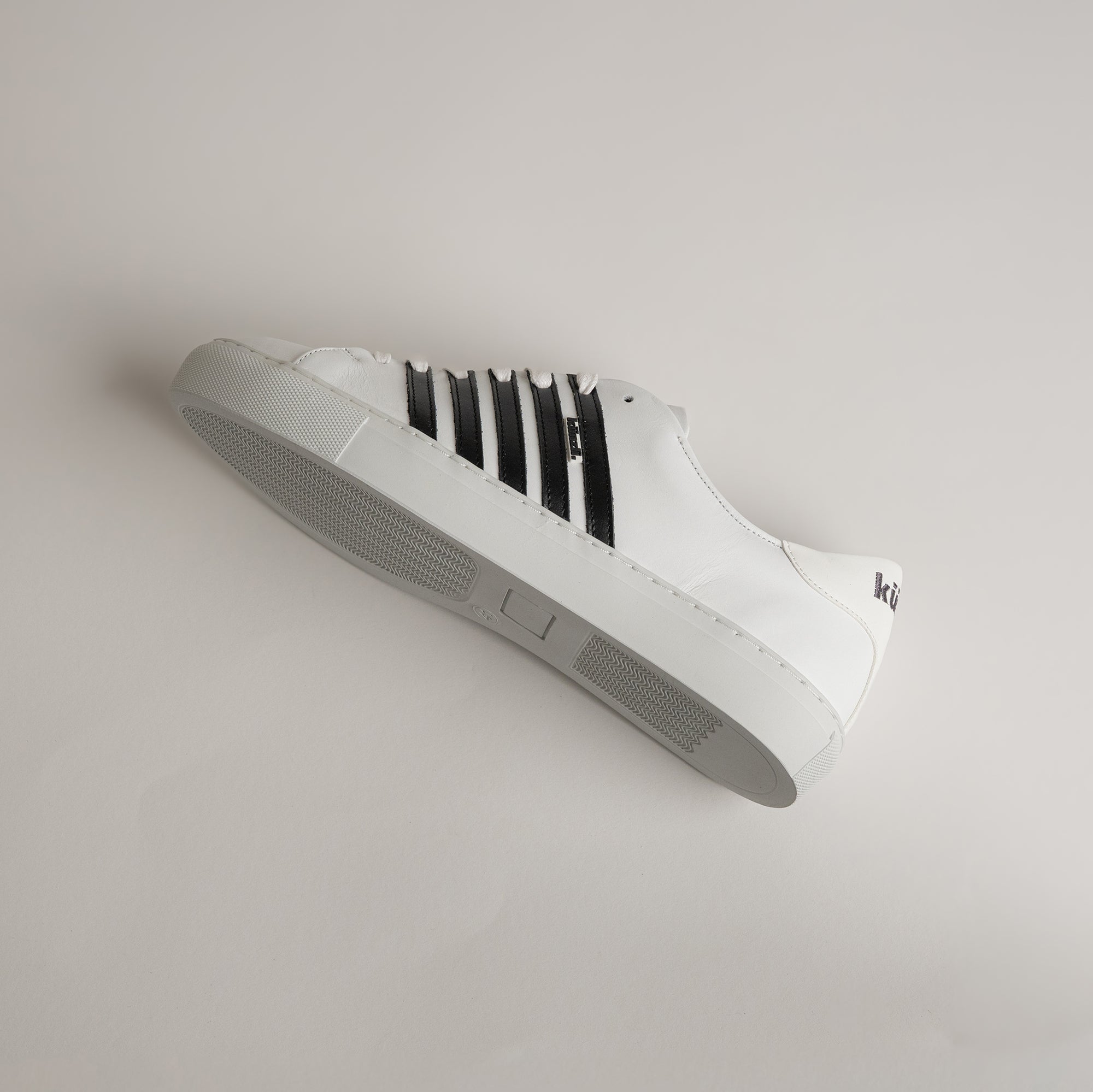 Weißer Kuenzli Schuh mit schwarzen Streifen auf hellem Hintergrund, seitlich liegend.