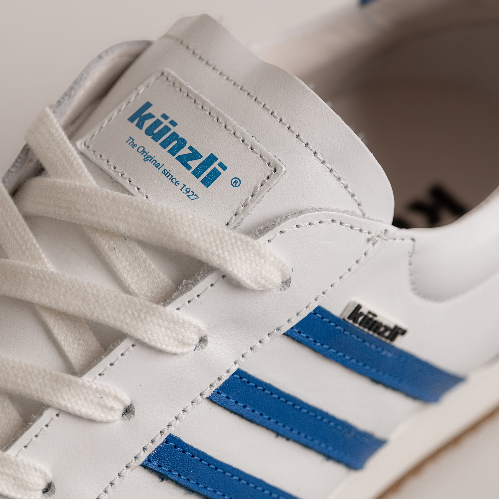 Weißer Kuenzli Schuh mit blauen Streifen und Logo, aus Leder gefertigt.