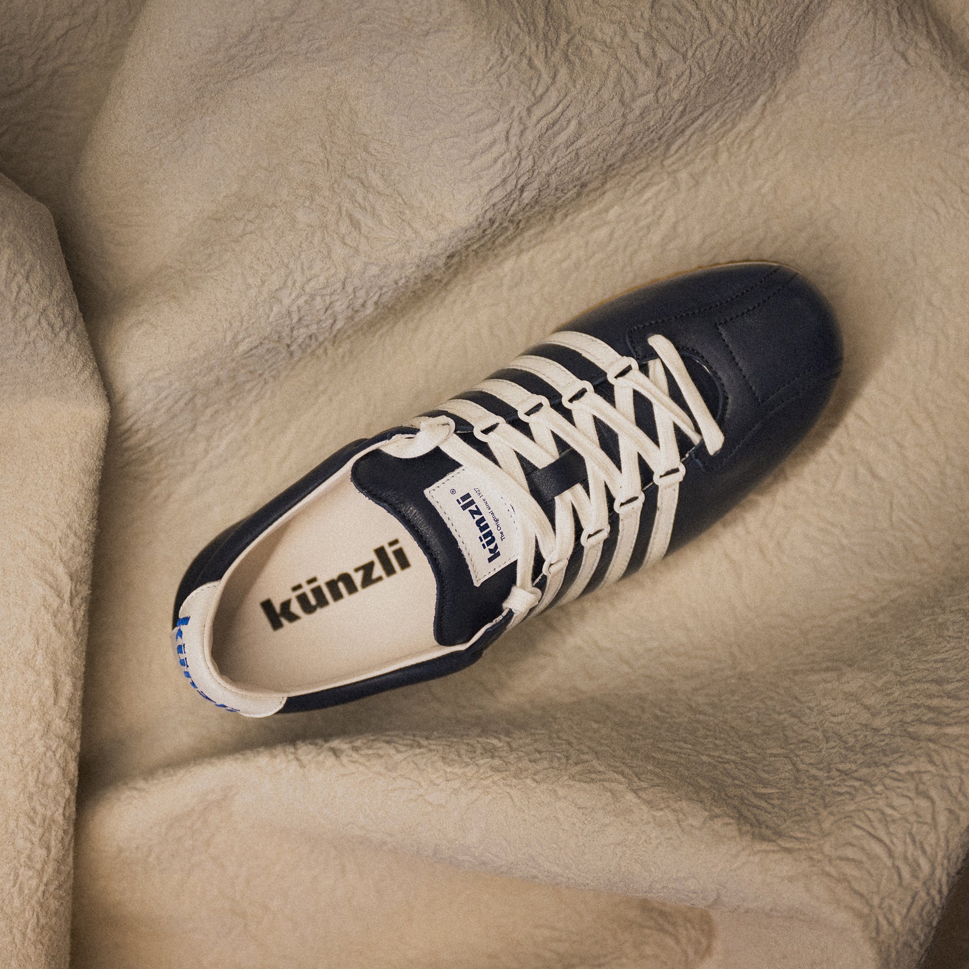 Schwarzer Kuenzli Schuh auf beigem Stoff, mit weißen Schnürsenkeln und Logo im Inneren.