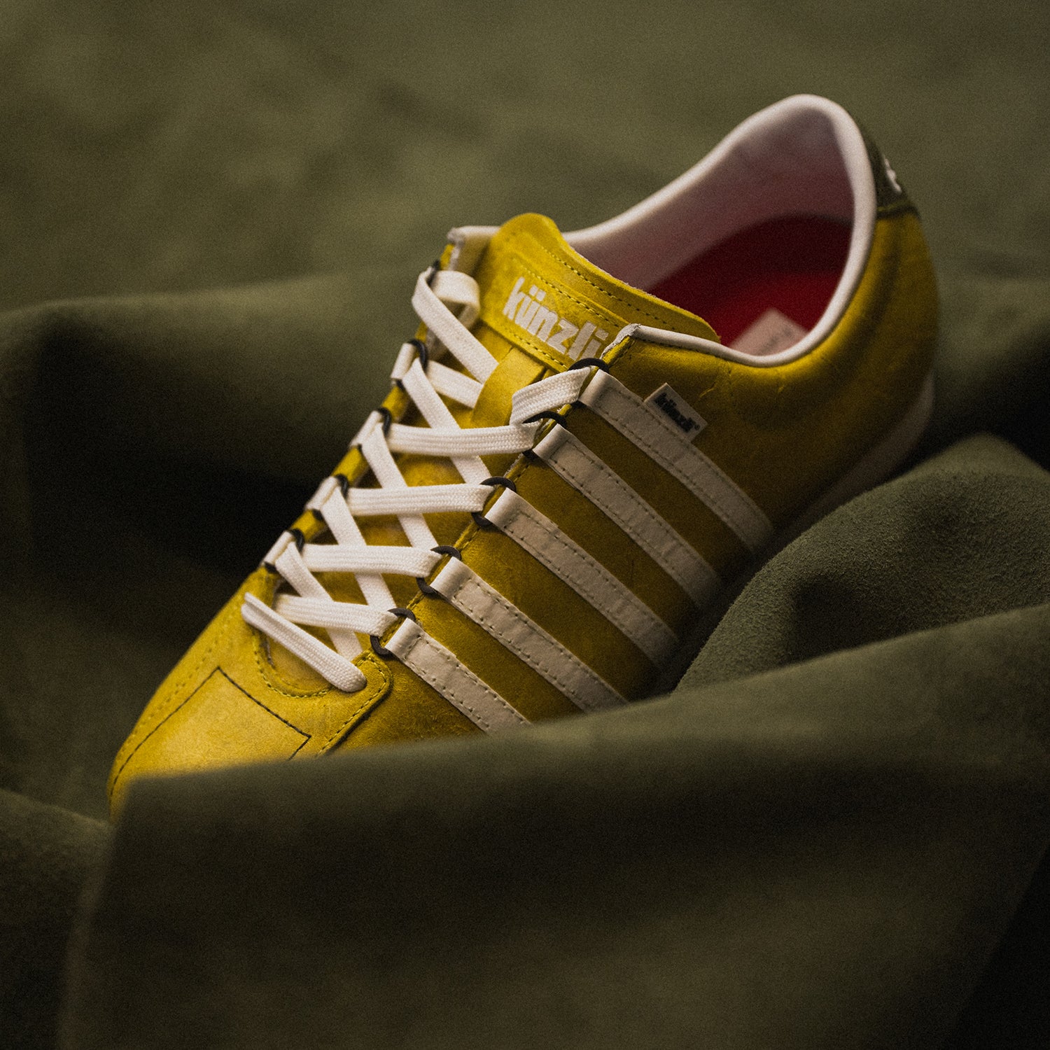 Gelber Kuenzli Schuh aus Leder mit weißen Streifen auf grünem Stoffhintergrund.