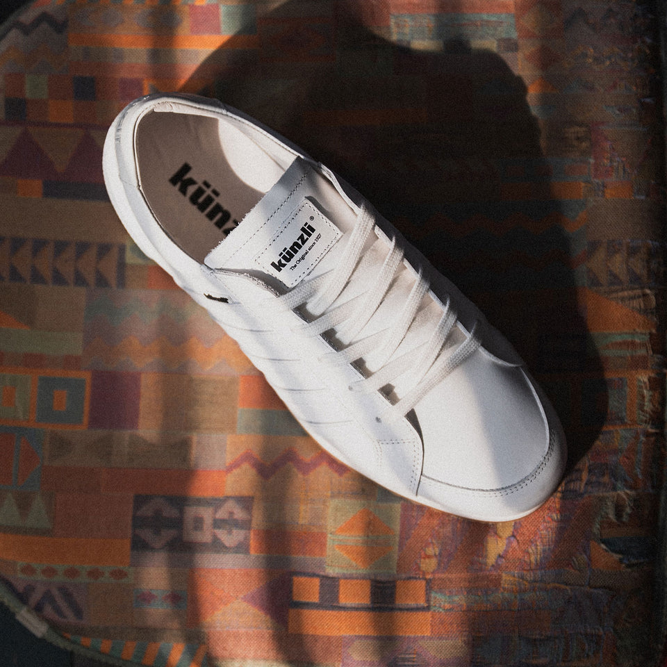 Ein weißer Kuenzli Schuh auf gemustertem Stoff, beleuchtet von Sonnenlicht.