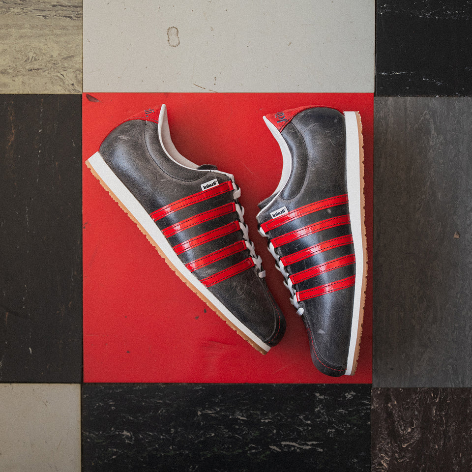 Graue Kuenzli Schuhe mit roten Streifen auf rotem und schwarzem Boden fotografiert.