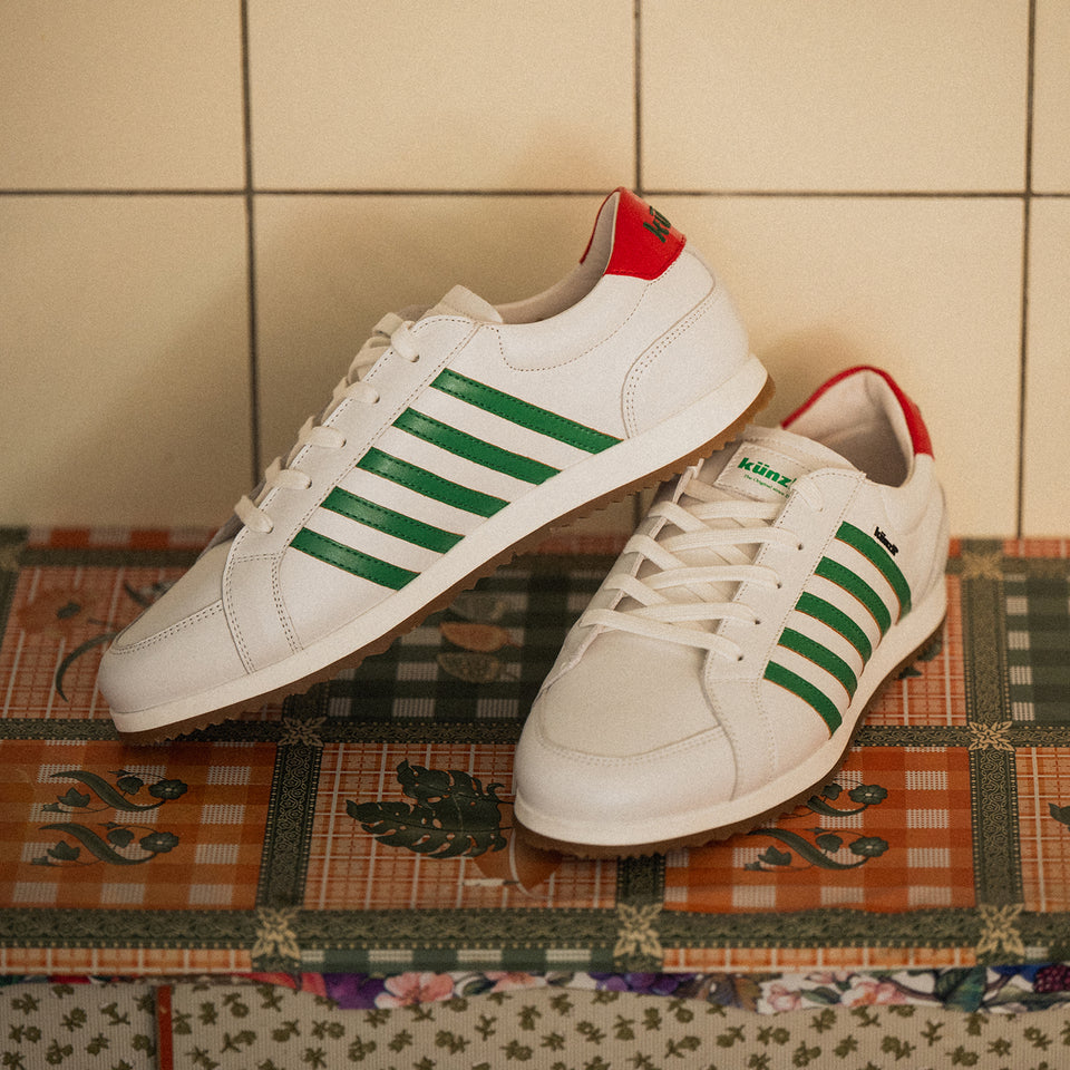 Weiße Kuenzli Schuhe mit grünen Streifen und rotem Detail auf gemustertem Untergrund.