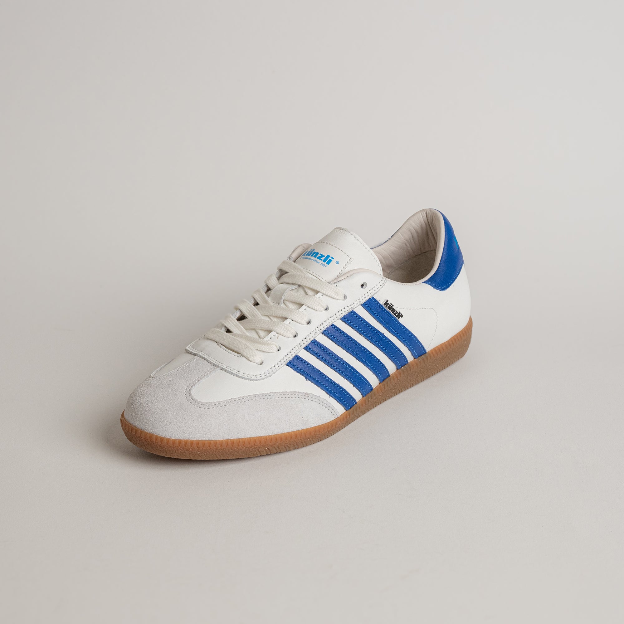 Weißer Kuenzli Schuh mit blauen Streifen und brauner Sohle auf hellem Hintergrund.