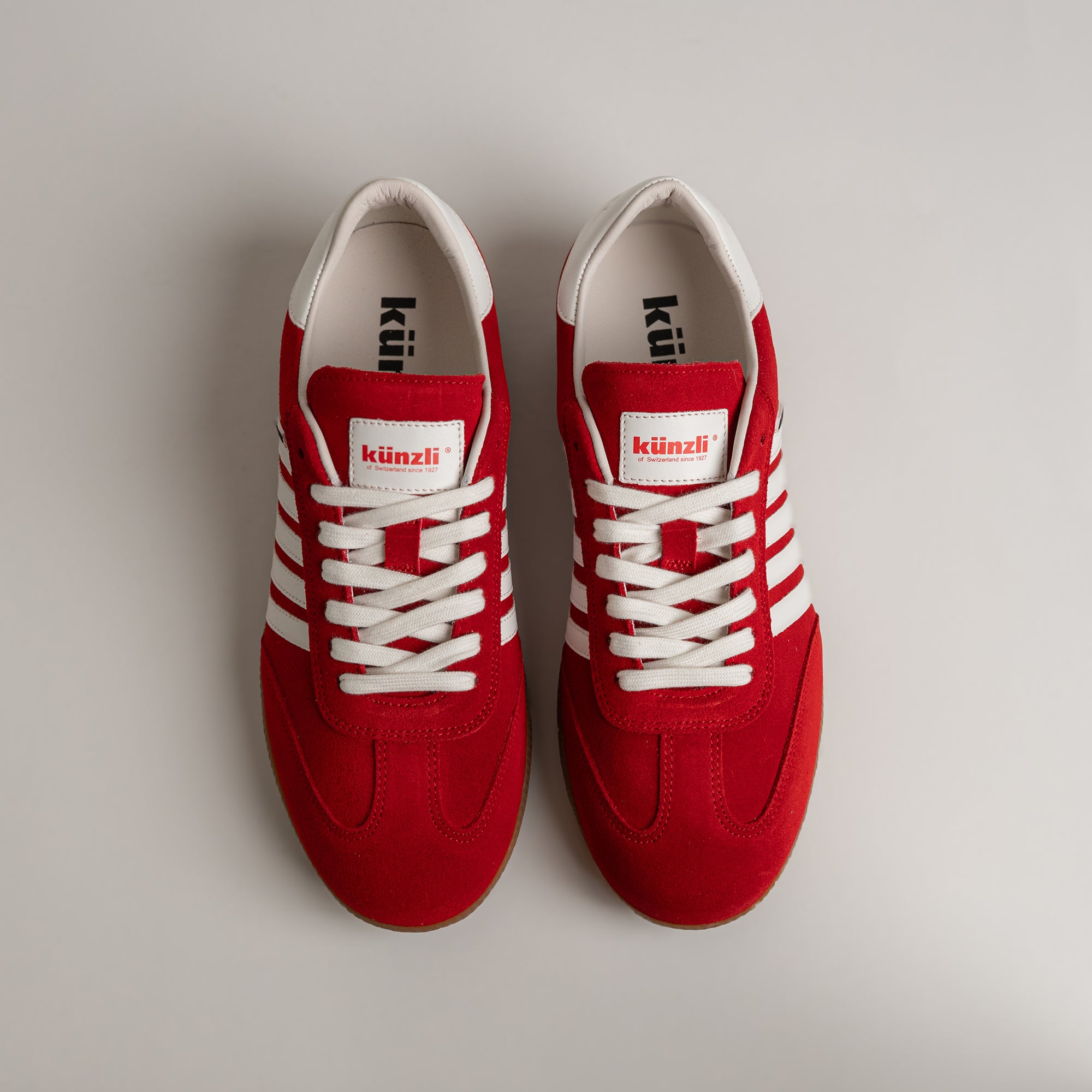 Rote Kuenzli Schuhe aus Leder mit weißen Streifen und Schnürsenkeln auf grauem Hintergrund.