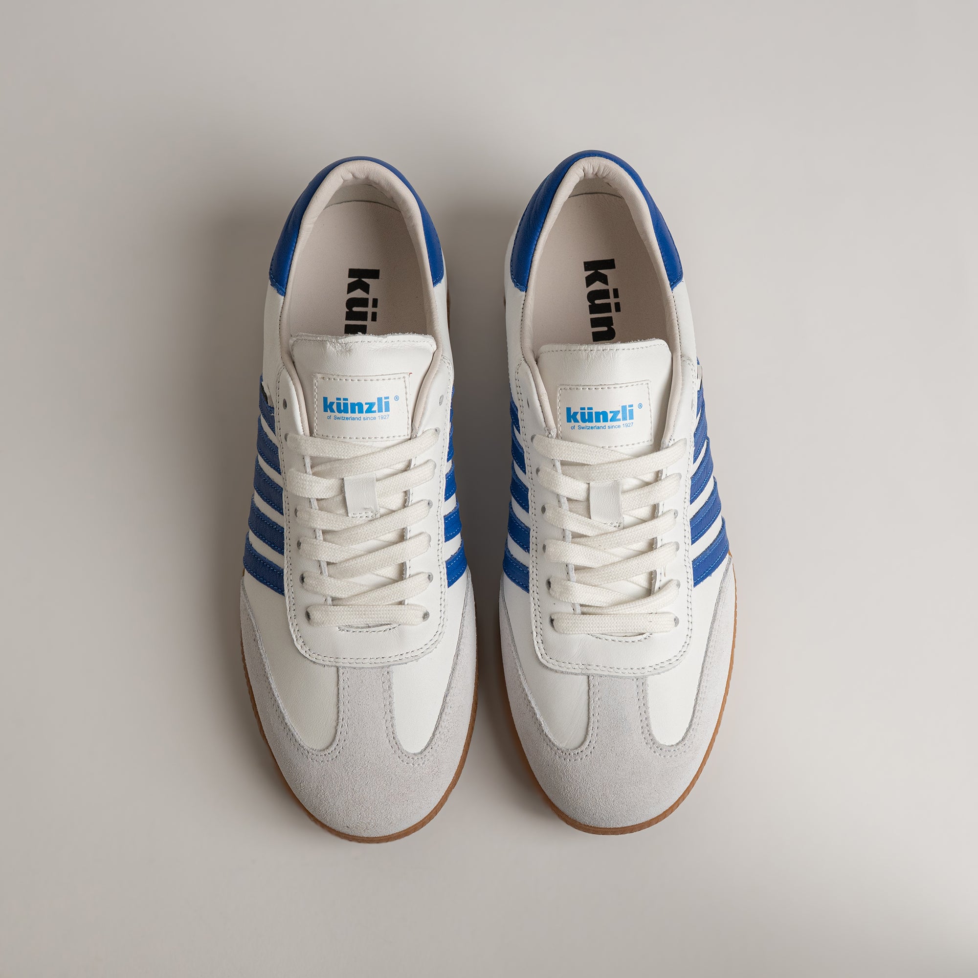 Weiße Kuenzli Schuhe mit blauen Streifen auf hellem Hintergrund, sportliches Design.
