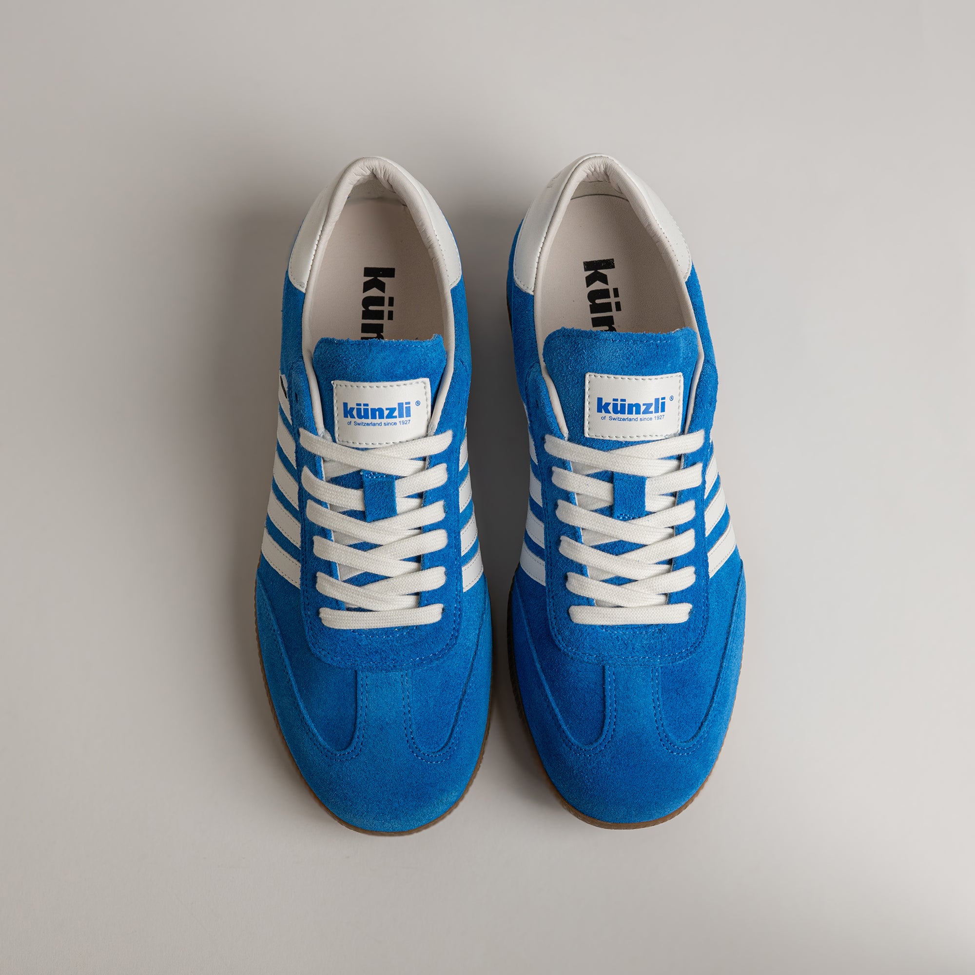 Blaue Kuenzli Schuhe aus Wildleder mit weißen Streifen auf grauem Hintergrund.