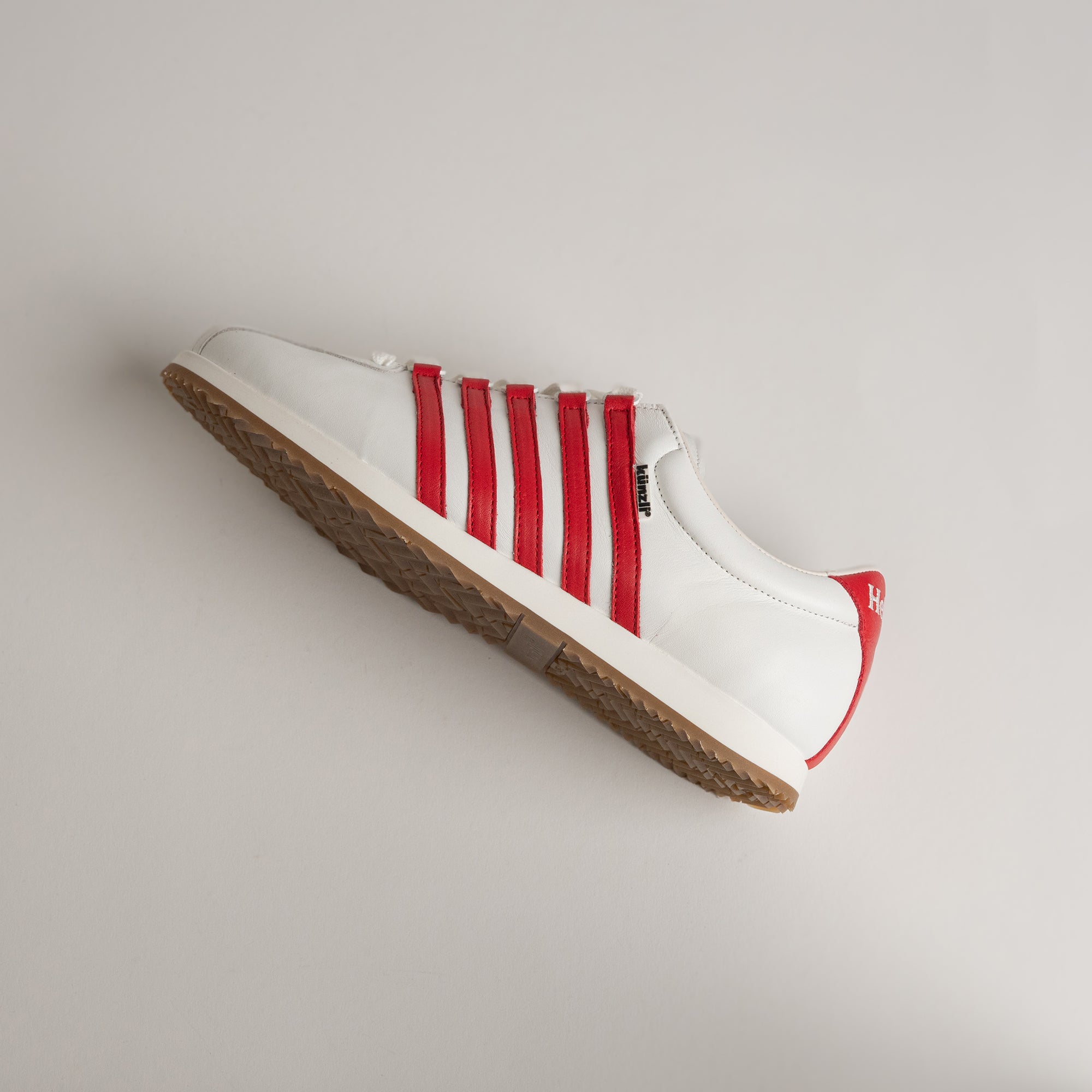 Weißer Kuenzli Schuh mit roten Streifen und brauner Sohle auf grauem Hintergrund.