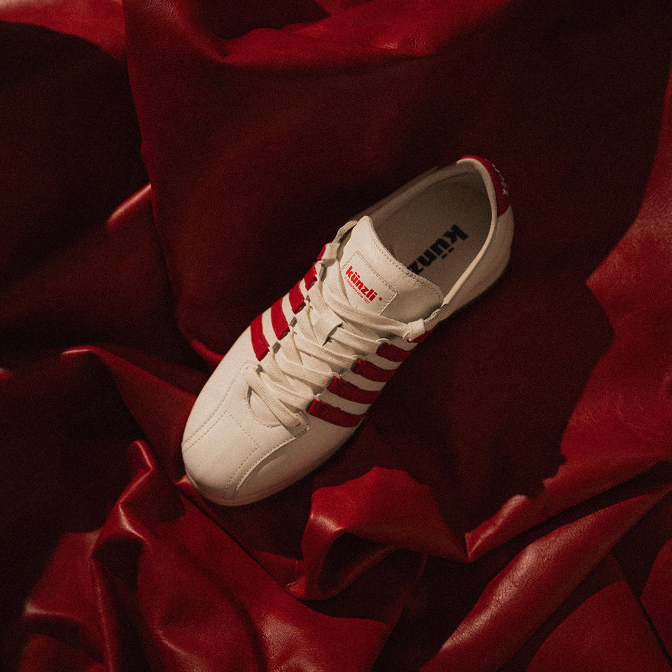 Ein weißer Kuenzli Schuh mit roten Streifen liegt auf rotem Leder.