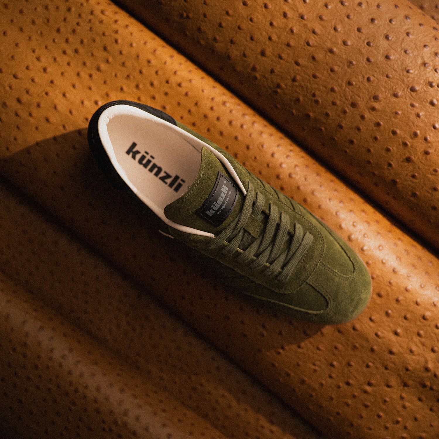 Grüner Kuenzli Schuh aus Wildleder auf braunem, strukturiertem Ledersessel präsentiert.