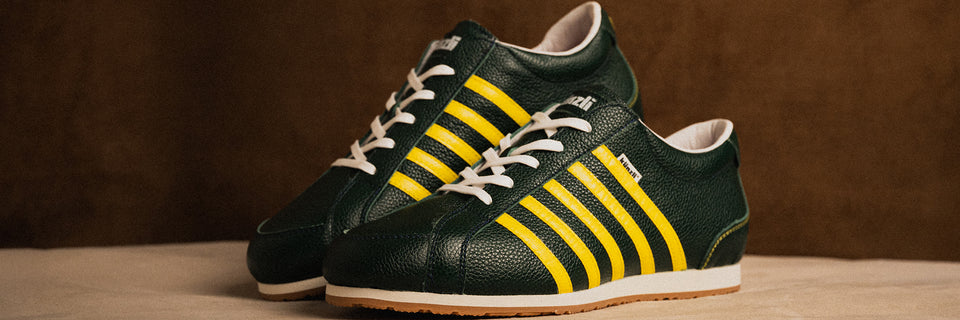 Grüne Kuenzli Schuhe mit gelben Streifen auf beigem Hintergrund präsentiert.