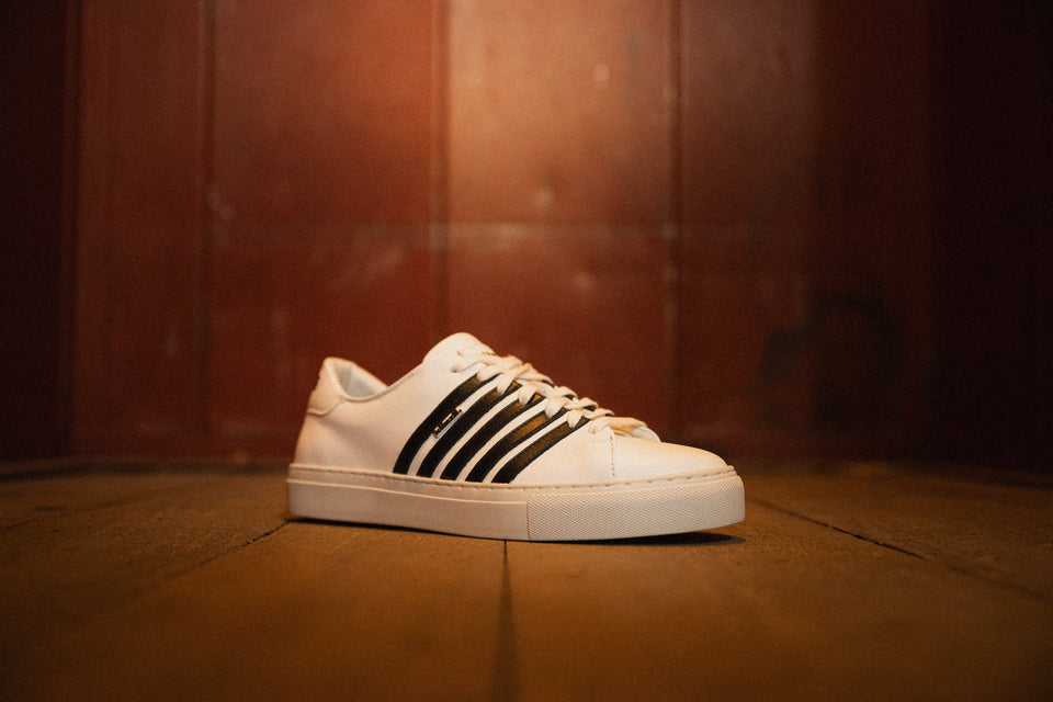 Weißer Sneaker von Kuenzli Schuhe mit schwarzen Streifen auf Holzboden präsentiert.