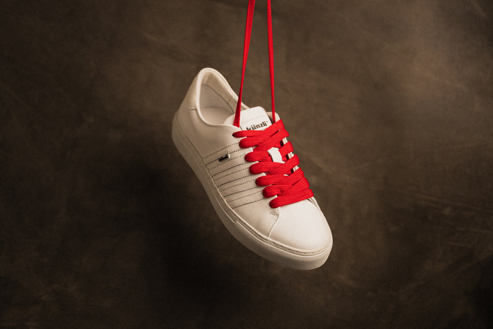 Ein weißer Kuenzli Schuh mit roten Schnürsenkeln hängt vor braunem Hintergrund.