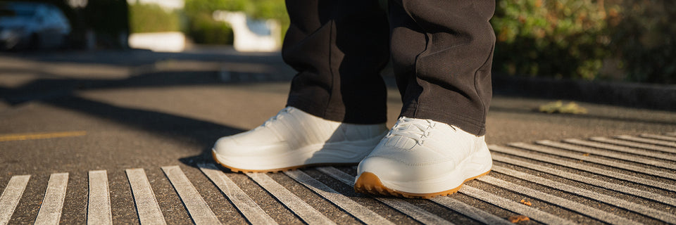 Person trägt weiße Kuenzli Schuhe auf Asphalt bei Sonnenschein.